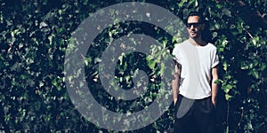 Bearded man with tattoo wearing blank white tshirt and black sunglasses.Green garden wall background. Wide mockup