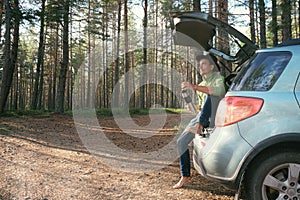 Bearded man tourist in felt hat with hot tea in thermos sitting in car trunk in pine forest. Road trip, weekend on nature, outdoor