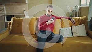 A bearded man is using mobile phone and making a deal sitting at home, freelance. 4K footage