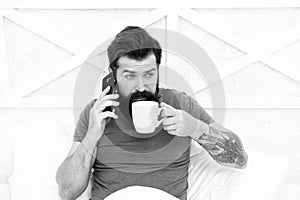 Bearded man using mobile technology in bed. Handsome guy talking on phone and drinking coffee at home. Modern life new