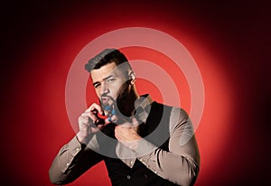Bearded man in vest on red background. Men cut their beard with hairdressing scissors