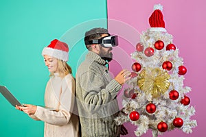 Bearded man in VR headset and pretty girl in Santa hat with laptop near Christmas tree. Modern technology and