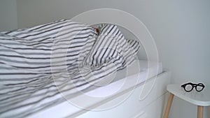 Bearded man waking up in bed under blanket, smiling, putting his eyeglasses on and smack