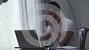 Bearded man in white t-shirt working with laptop and papers drinking coffee at home or in the office. The guy is tired