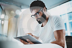 Bearded man in white tshirt wearing eye glasses and using portable electronic pro tablet computer at modern lightful