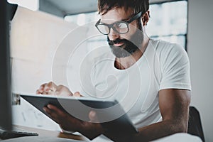 Bearded man in white tshirt working with portable electronic pro tablet computer at modern lightful office.Horizontal