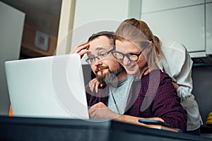 Bearded man and woman with eyeglasses using laptop in the home interior, communicate with friends online, laugh and grimace