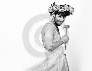 Bearded man in a woman`s wedding dress on her naked body, holding a flower. on his head a wreath of flowers. funny