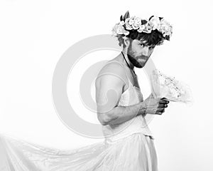 Bearded man in a woman`s wedding dress on her naked body, holding a flower. on his head a wreath of flowers. funny