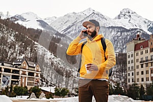 Bearded man in yellow hoodie with backpack stands on background of high snowy mountains, talking on mobile phone, drinking coffee