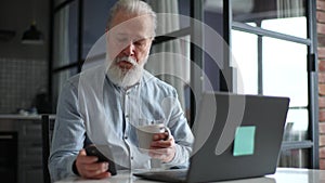 Bearded mature elderly man with grey hair dialing up phone number for online shopping looking on laptop screen, sitting