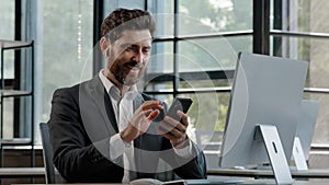 Bearded middle-aged business man in office sitting table using phone mobile audio app service to listening music singing
