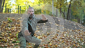 Bearded middle-aged man doing chinese tai chi exercises on lawn in autumn city park