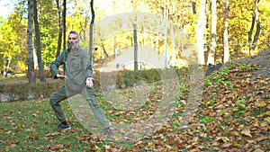 Bearded middle-aged man in khaki clothes doing chinese tai chi exercises on lawn in autumn city park