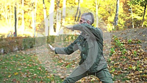 Bearded middle-aged man in khaki clothes doing chinese tai chi exercises on lawn in autumn city park