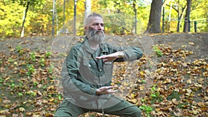 Bearded middle-aged man in khaki clothes doing chinese tai chi exercises on lawn in autumn city park