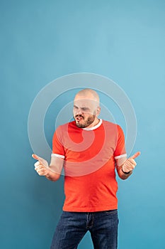 A bearded middle-aged man in a red T-shirt on a blue background rejoices and shows his thumbs up. Isolated
