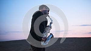 bearded musician in sunglasses and black jacket performs his part on beautiful shiny saxophone at sunset or sunrise with