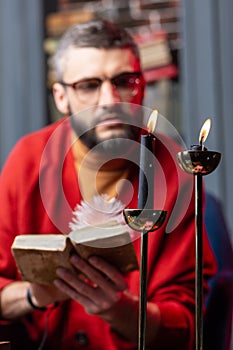 Bearded mysterious diviner reading book near flaming candles