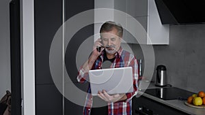 Bearded old man using laptop in kitchen and talking by phone at the same time