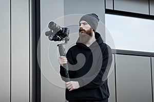 Bearded Professional videographer in black hoodie holding professional camera on 3-axis gimbal stabilizer. Filmmaker