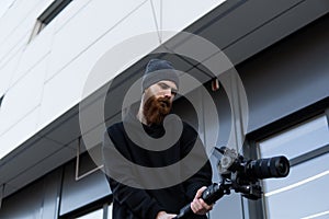 Bearded Professional videographer in black hoodie holding professional camera on 3-axis gimbal stabilizer. Filmmaker