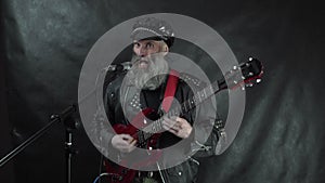 Bearded rock musician in leather jacket and cap plays red electric guitar and sings on stage of nightclub concert black