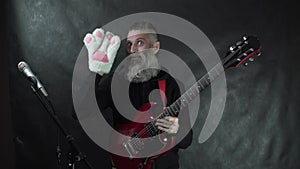 Bearded rockstar with black hoodie, red electric guitar and shaved temples playing with white cat paw glove on nightclub