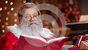 Bearded Santa Claus reading magic book with childish Christmas wishes at glowing room home interior