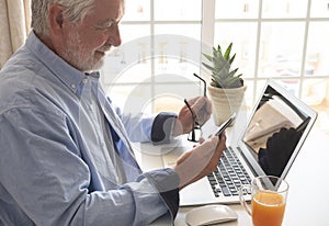 Bearded senior man stays at home using cellphone. Desk office with laptop computer and orange juice glass.Seniors and technology