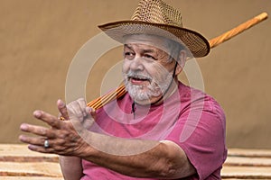 Man in straw hat sitting against clay wall, gesticulating with left hand while  holding wicker walking stick in right one o