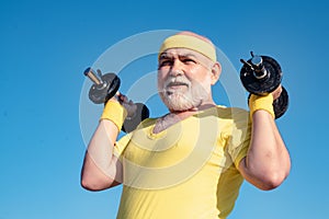 Bearded Senior sportman exercising with lifting dumbbell on blue sky background. Isolated, copy space. Senior man