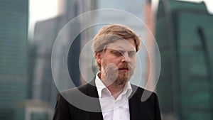 A bearded, serious man in a jacket and shirt is in the open air against the background of the business center of Moscow