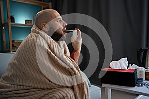 Bearded sick caucasian man in blanket using oral spray at home. Rhinitis, medicine and healthcare