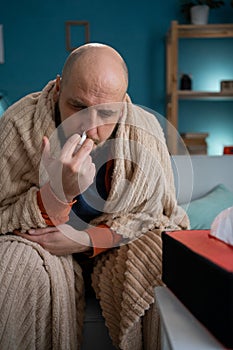 Bearded sick man with Cold Sitting on Sofa in Blanket at Home, using nasal spray. Sick male person. Disease, Healthcare