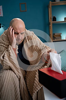 bearded sick man on couch at home suffers from runny nose, sneezes. Unwell guy taking napkin feeling bad fever virus