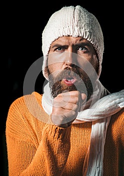 Bearded sick man in sweater, hat and scarf coughing. Handsome guy having sore throat and coughing into his fist