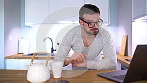 Bearded sleepy man having breakfast in the kitchen and using a laptop