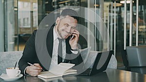 Bearded smiling businessman using laptop computer, talking on smartphone and writing down business information in cafe