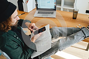 Bearded student sits with his feet up on his desk while studying online using laptop and smartphone