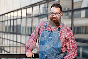 Bearded stylish craftsman wearing blue overalls looking at camera, outdoors. Family workshop, old manufactory worker