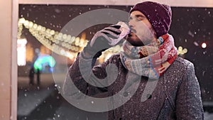 Bearded stylish man in gray coat and knitted scarf drinks coffee out of paper cup and looks to the camera smiling