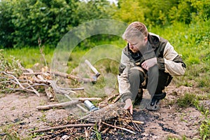 Bearded survivalist male in raincoat putting brushwood on campfire to making fire in forest on cloudy summer day. photo