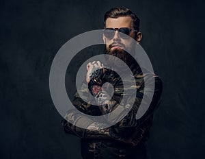 Bearded tattooed guy in military shirt and sunglasses posing with thoughtful look. Studio photo against dark wall