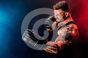 Bearded tattooed sportsman muay thai boxer in black undershirt and boxing gloves fighting on dark background with smoke.