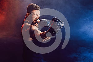 Bearded tattooed sportsman muay thai boxer in black undershirt and boxing gloves fighting on dark background with smoke.
