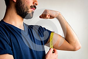 Bearded thin man measuring biceps, muscles of his left arm with a yellow tape measure. He`s calm, serious, quiet.