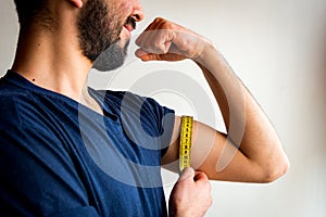 Bearded thin man measuring biceps, muscles of his left arm with a yellow tape measure. He`s smiling, happy.