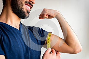 Bearded thin man measuring biceps, muscles of his left arm with a yellow tape measure. He`s smiling, happy.