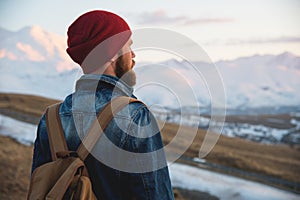 Bearded tourist hipster man in a hat with a backpack stand back on a roadside bump and watching the sunset against the